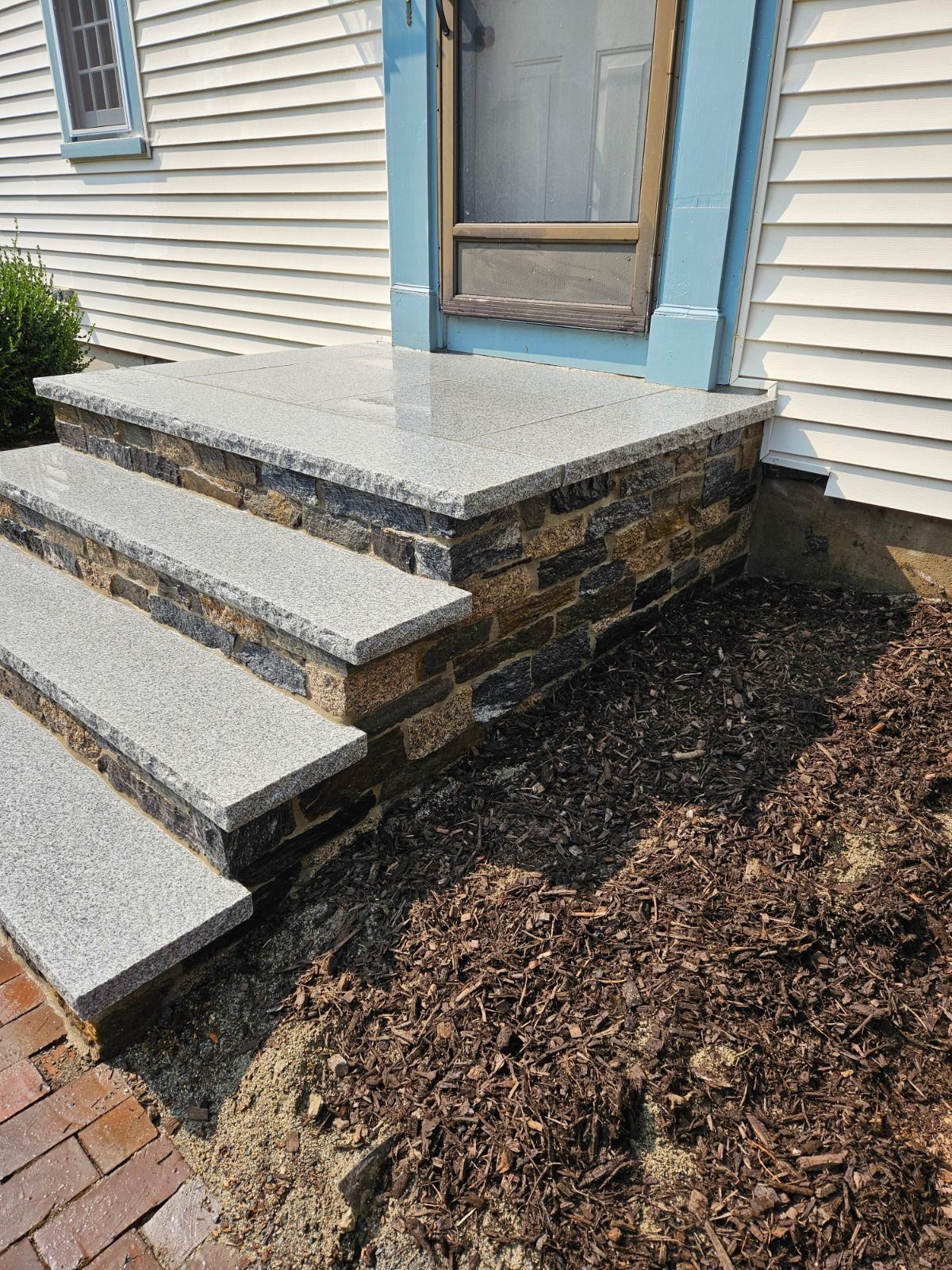 Front steps with a stone landing and brick base beside a house, next to a mulched garden bed.