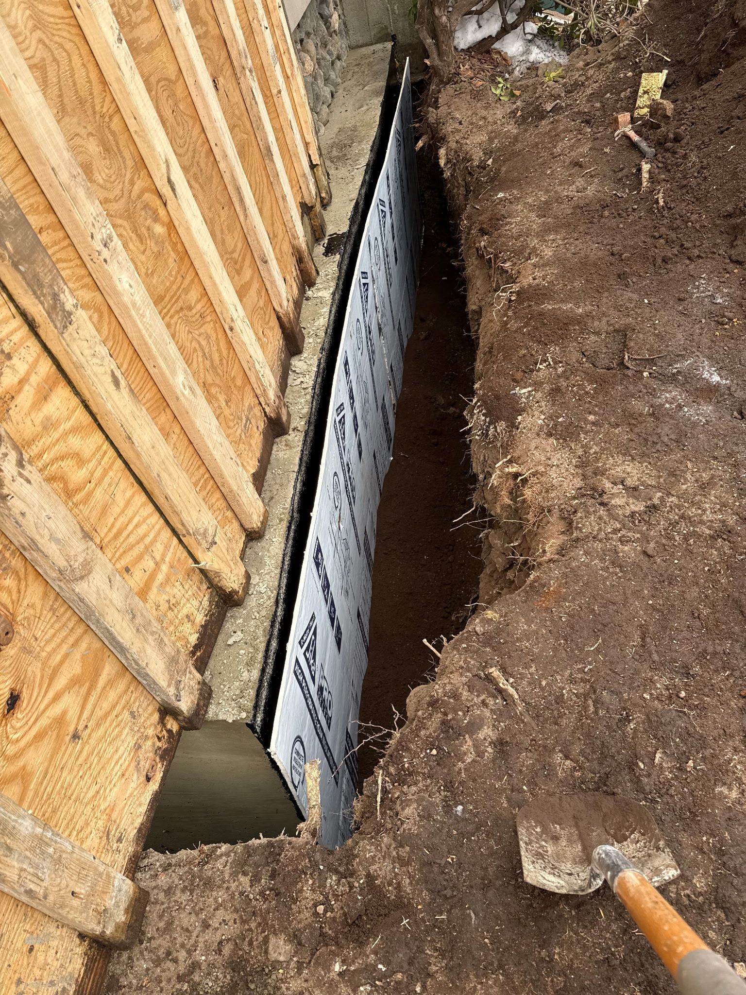 Trench beside a wooden fence with a metal foundation barrier and a shovel in dirt.