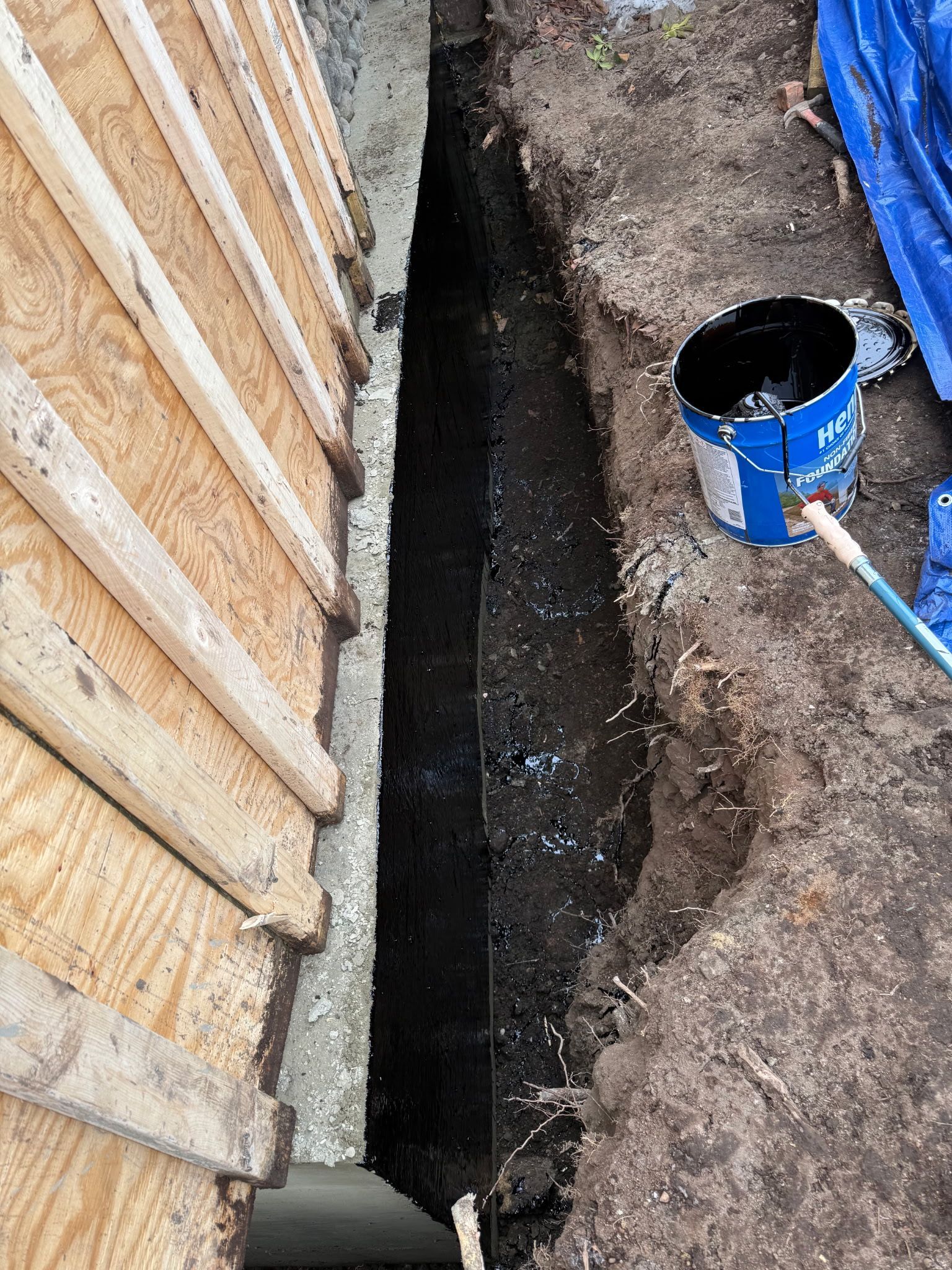 Narrow trench dug beside a wooden wall, with a blue bucket and shovel in the dirt.