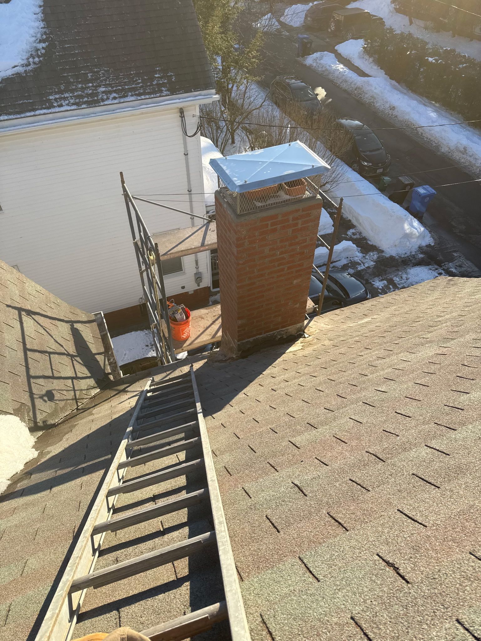 Rooftop ladder leading to a brick chimney beside a snowy roof and neighboring building.