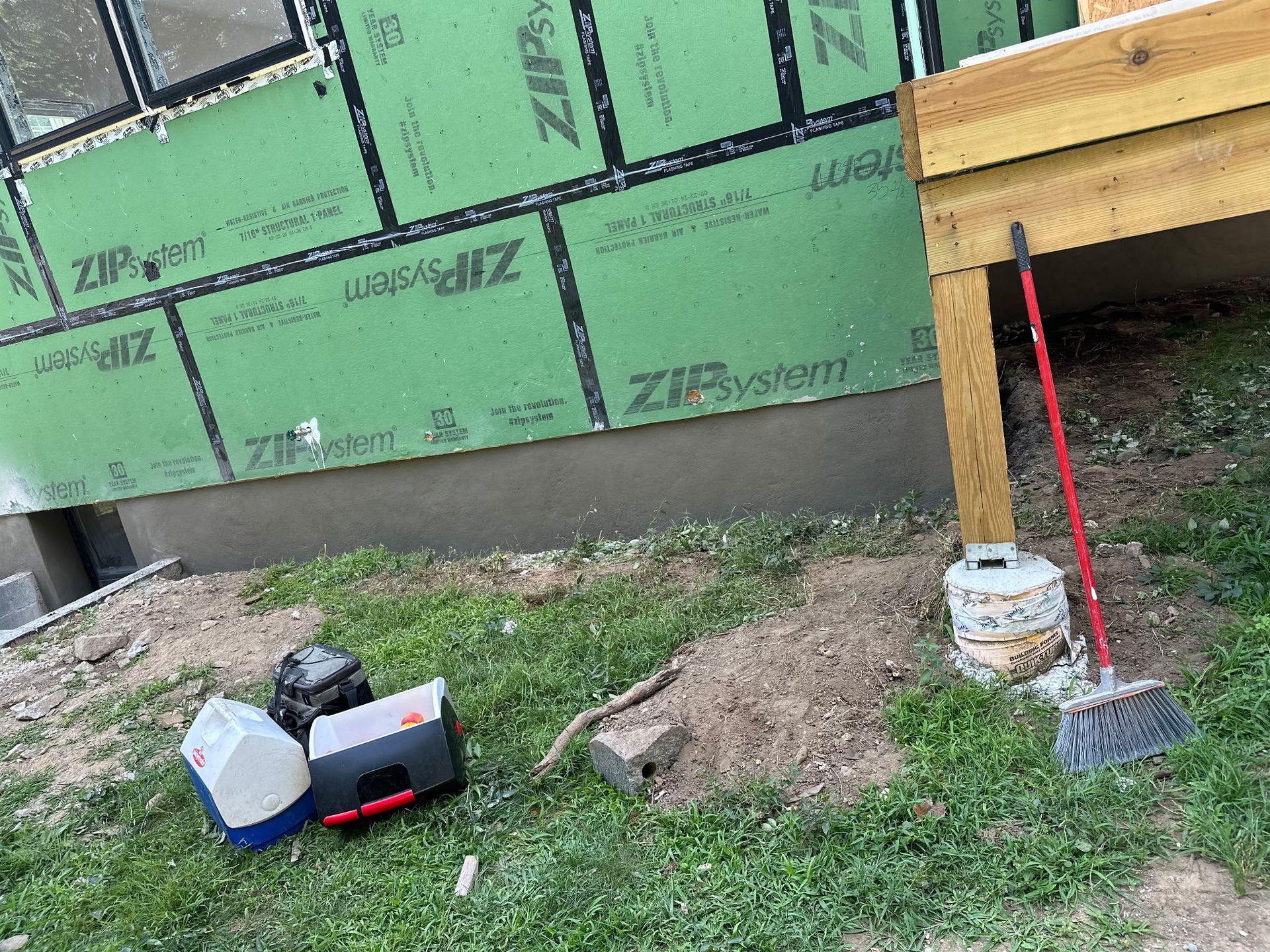 Partially built house exterior with green ZIP sheathing, wooden deck post, and tools on grass below
