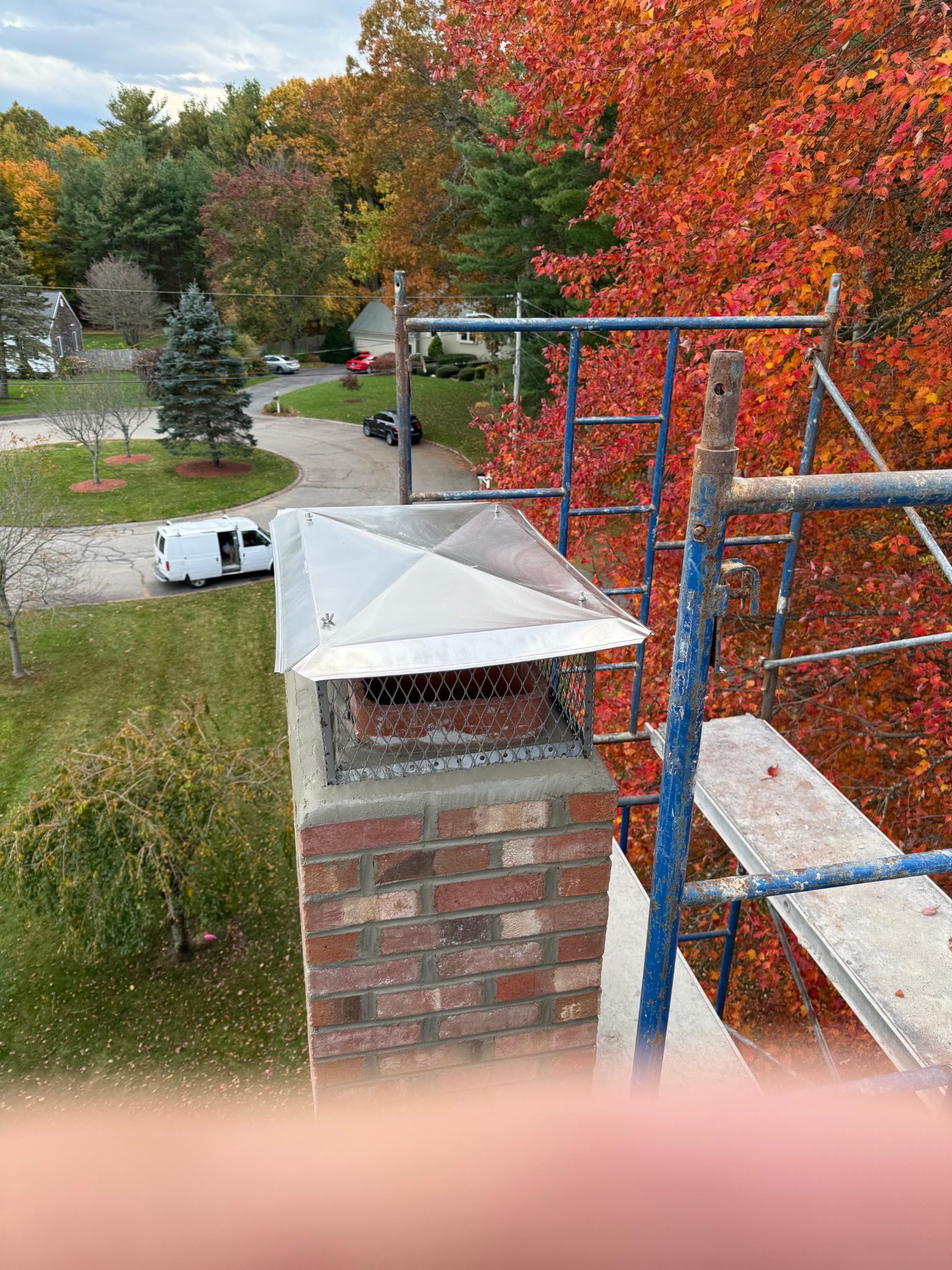 Brick chimney with metal cap beside a blue scaffold, with autumn trees and a white van below.
