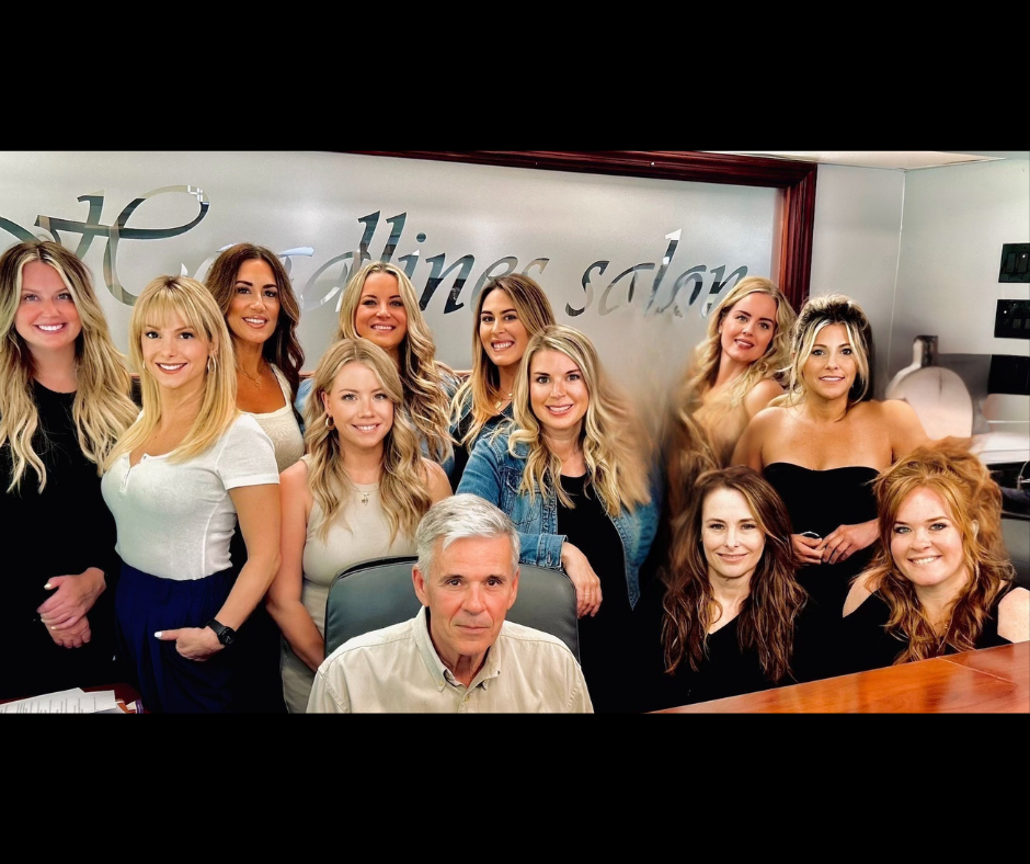 Group of people posing at a salon desk. A man is seated, with women surrounding him. The salon sign is visible.