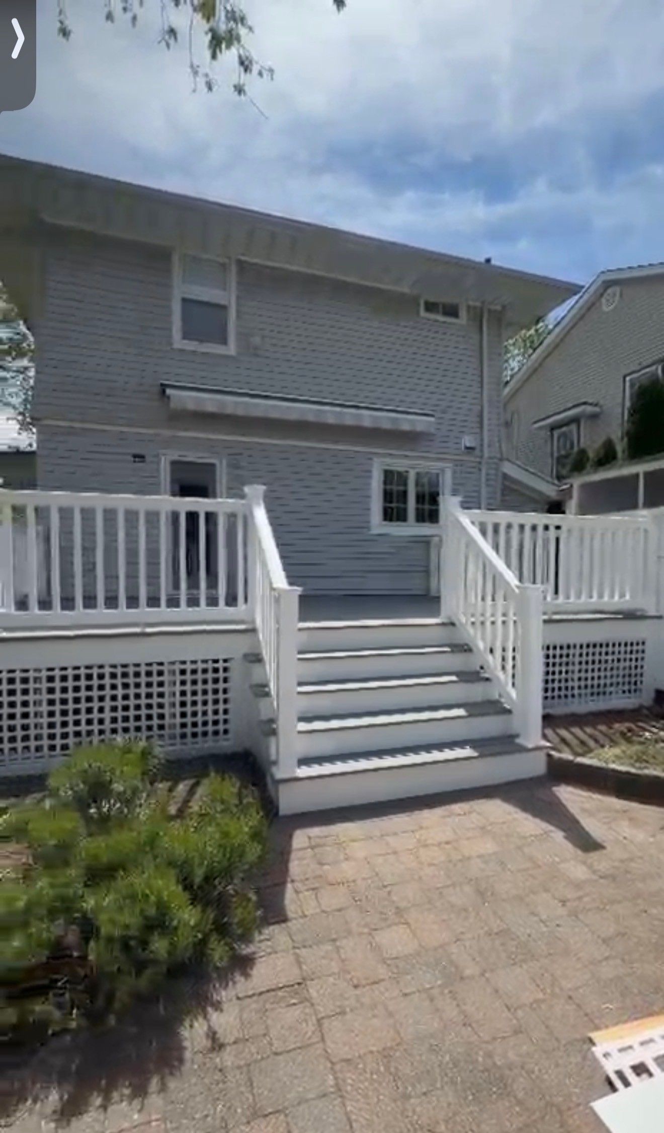 Gray two-story beach house with white railings, front stairs, and a sunny brick patio.