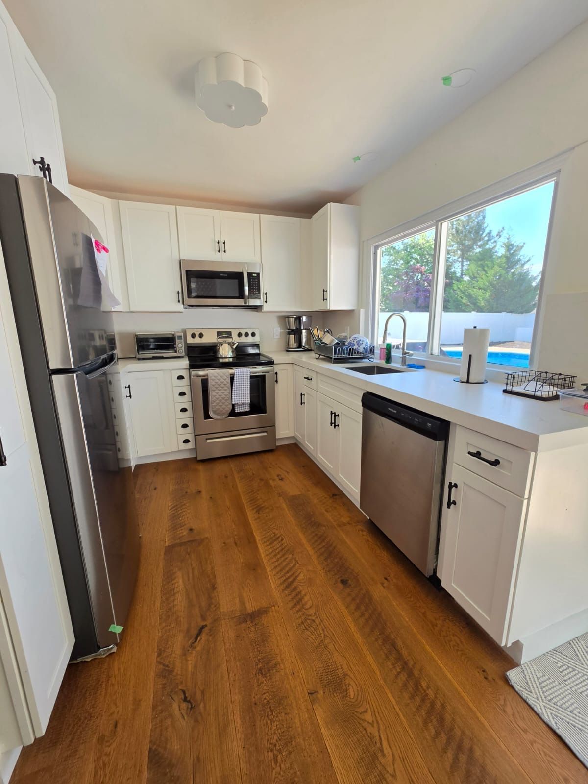 Bright modern kitchen with white cabinets, stainless appliances, wood floors, and a large window by the sink.