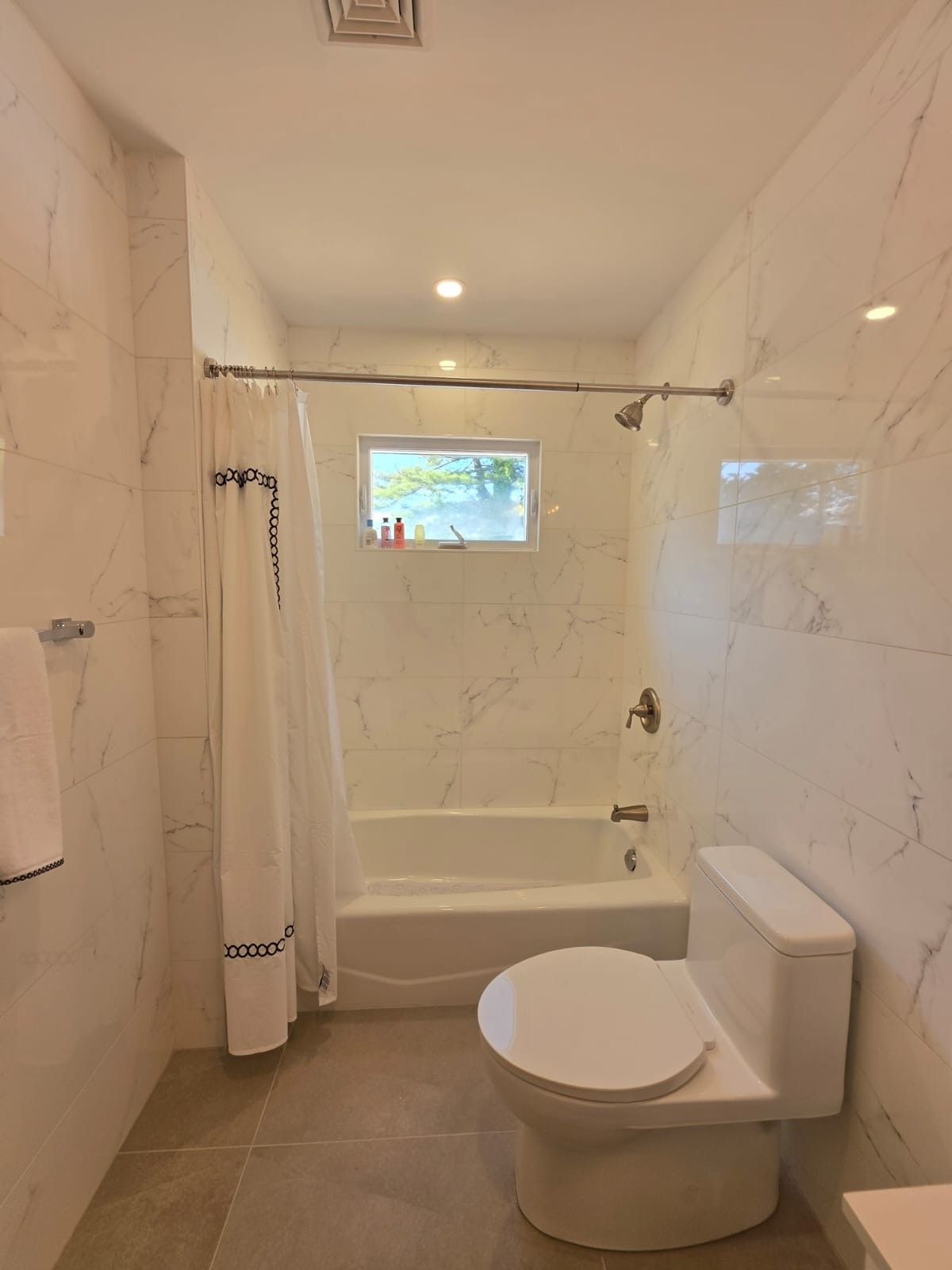 Modern bathroom with white tile, toilet, bathtub, and a small window above the tub