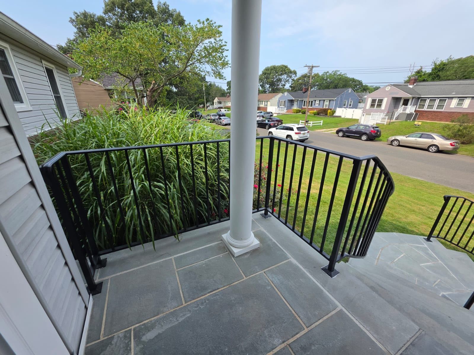 Front porch with black railing and gray stone floor, overlooking a suburban street and houses.