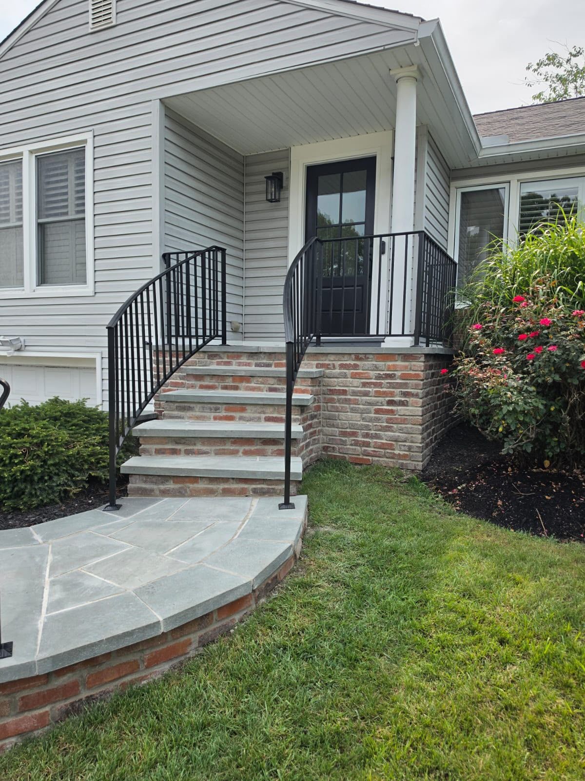 Front porch with brick steps, black railings, and a gray house exterior beside a lawn and shrubs