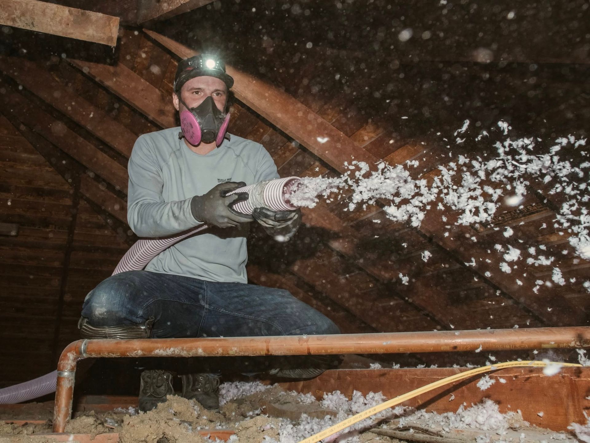 Person in attic sprays insulation; wearing respirator, headlamp, and gloves.