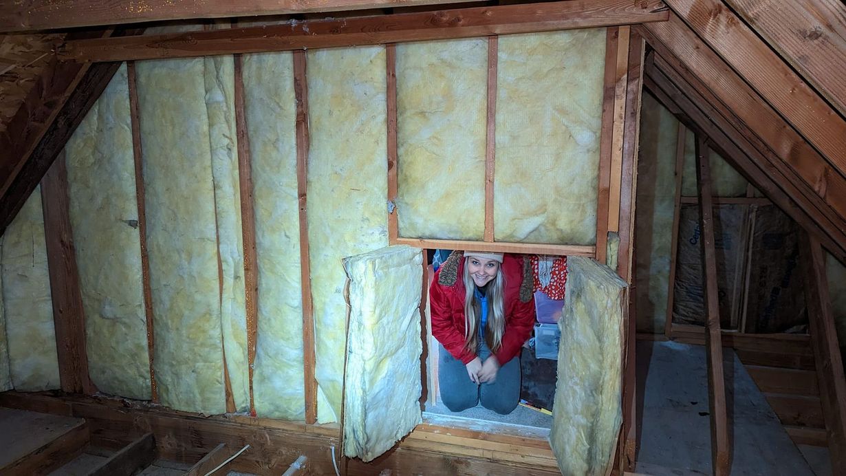 Woman kneeling in attic opening, surrounded by insulation. She&rsquo;s wearing a red jacket and smiling.