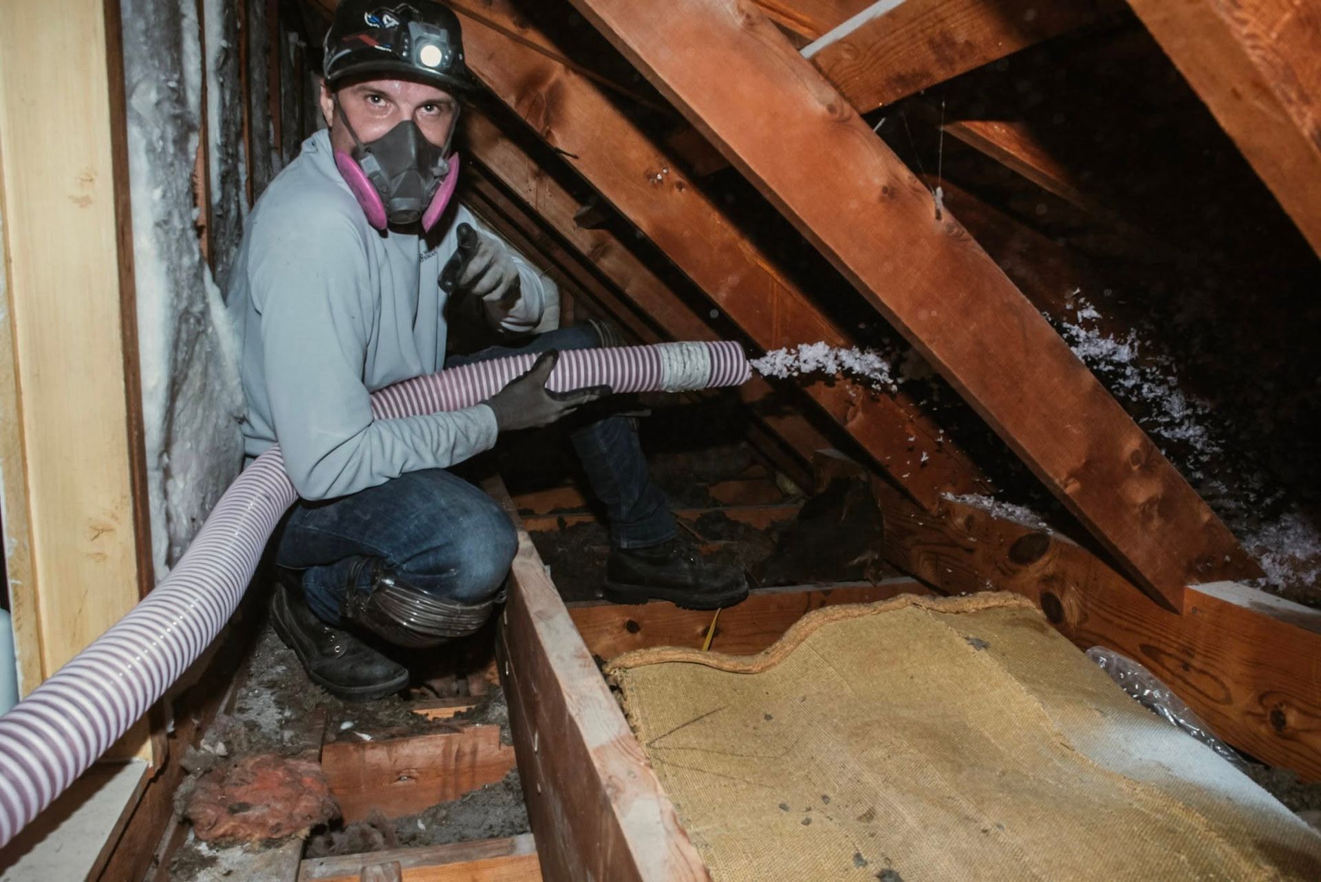 Man in attic wearing a respirator and gloves, blowing insulation. Brown wood beams and insulation fill the space.