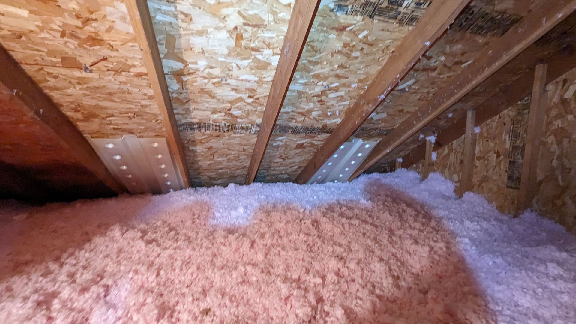Pink insulation filling an attic space, visible wooden beams and oriented strand board overhead.