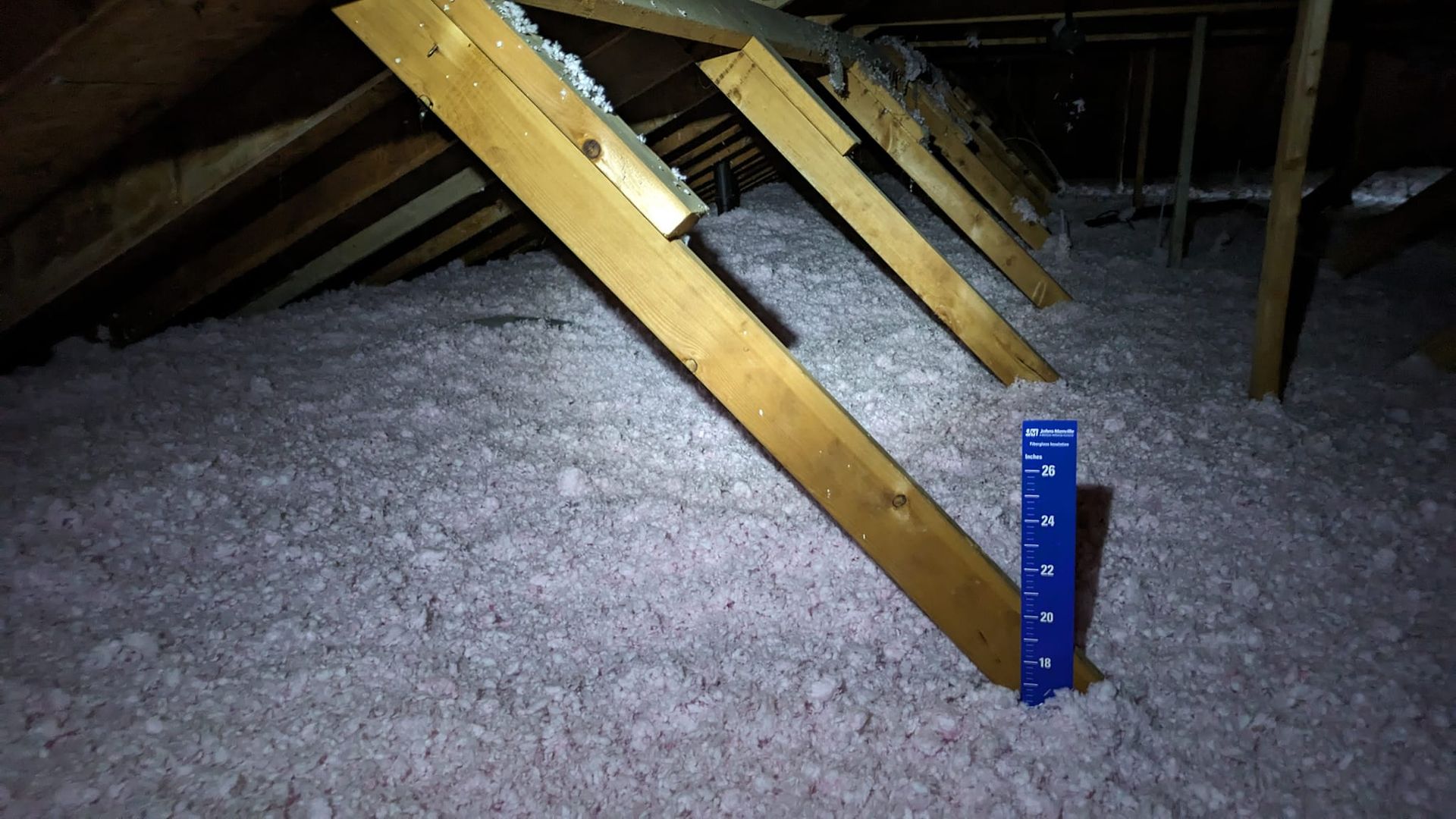 Attic with pink insulation and wooden beams; blue ruler shows insulation depth.
