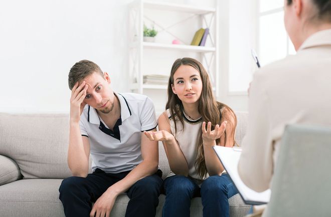 A couple sits on a couch in a counseling session, one person looking distressed while the other explains with gestures.
