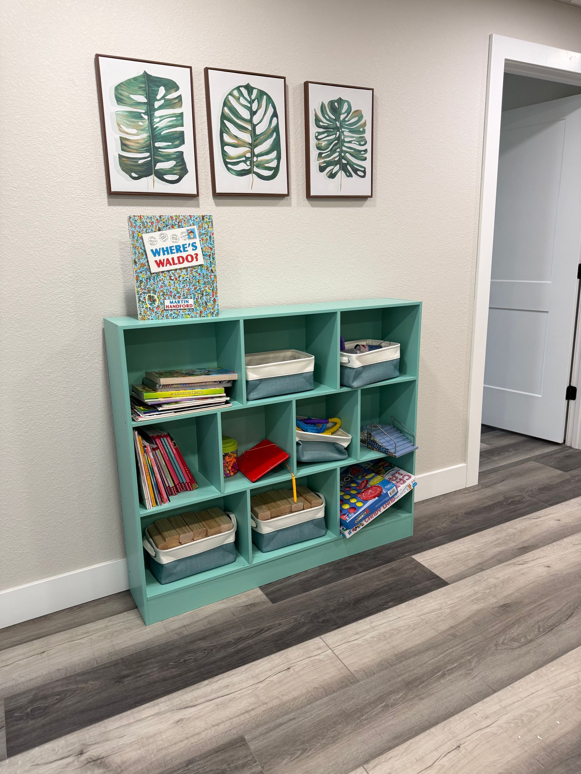 A light green cubby shelf filled with books and containers stands against a wall below three framed leaf art prints.