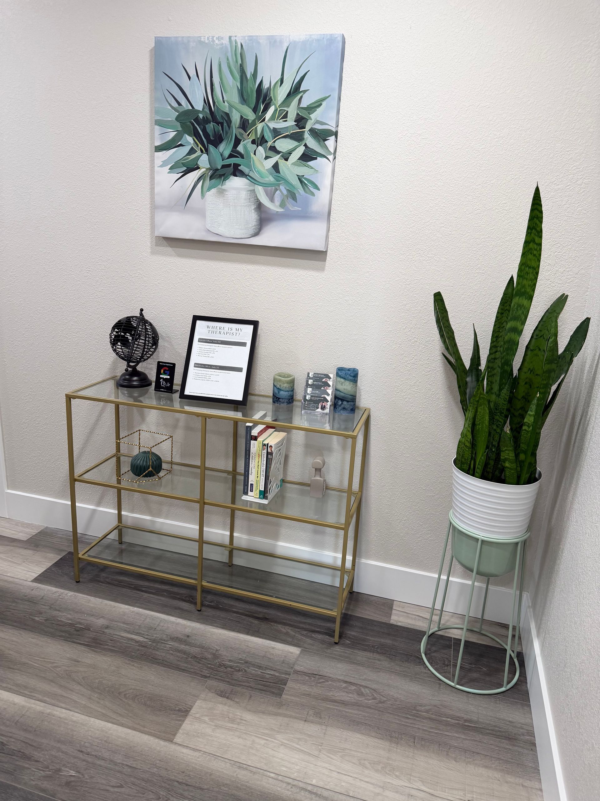 A gold-framed glass console table holds decor against a light wall, next to a potted snake plant on a metal stand.