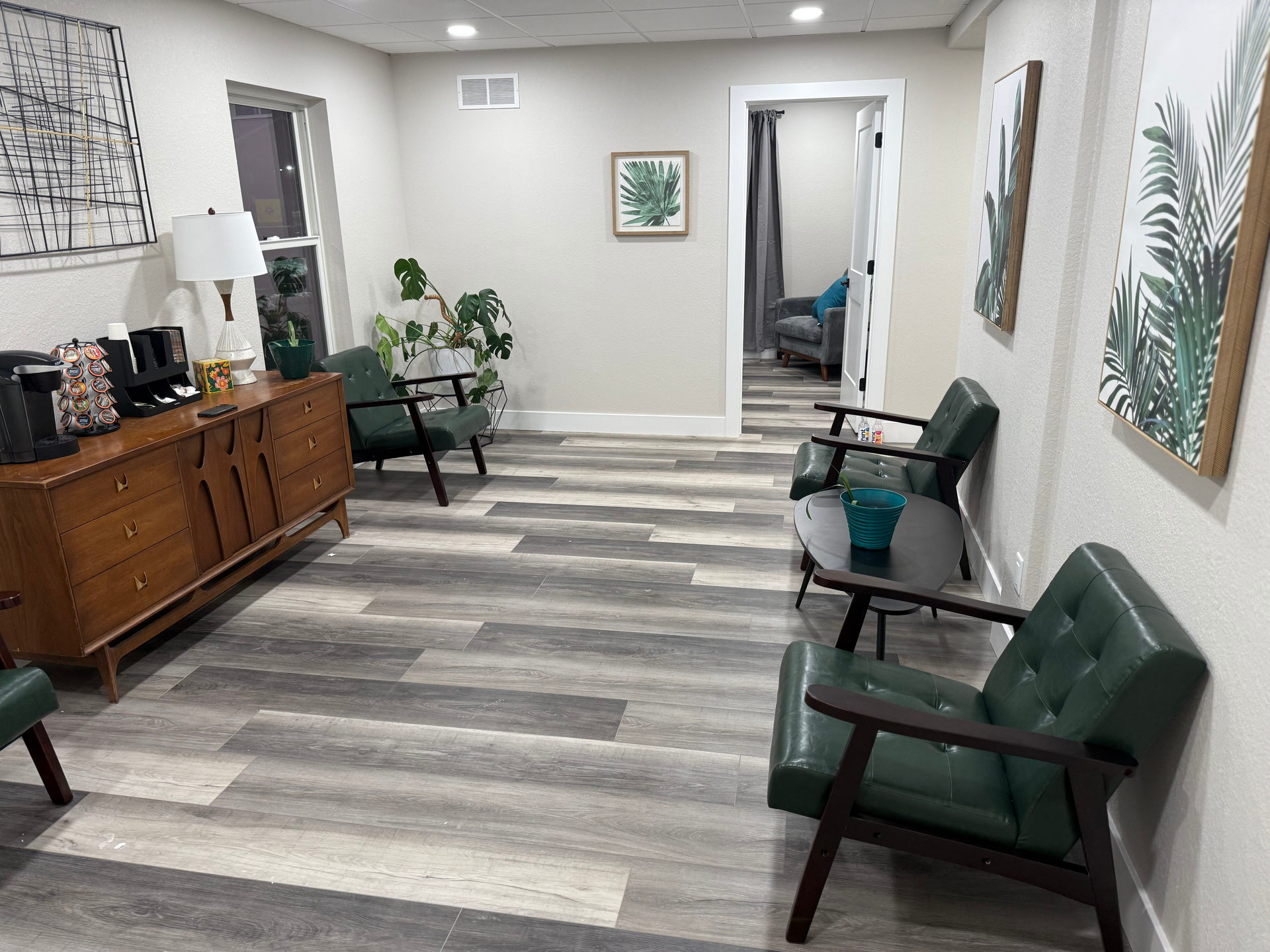 Waiting room with green chairs, wooden cabinet with coffee station, and plant art.