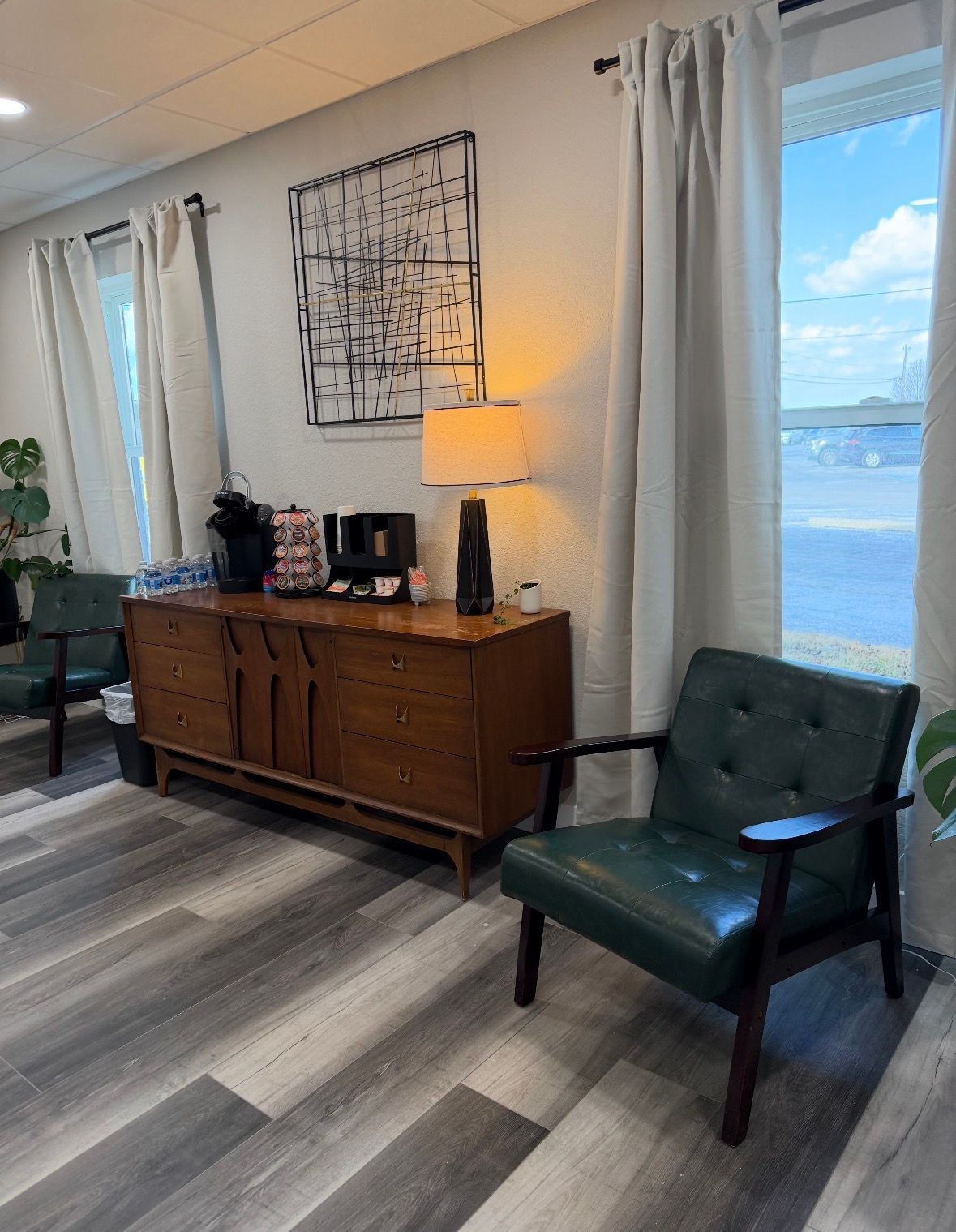 A room with a wooden sideboard, a lamp, wall art, and two green mid-century chairs placed by windows with light curtains.