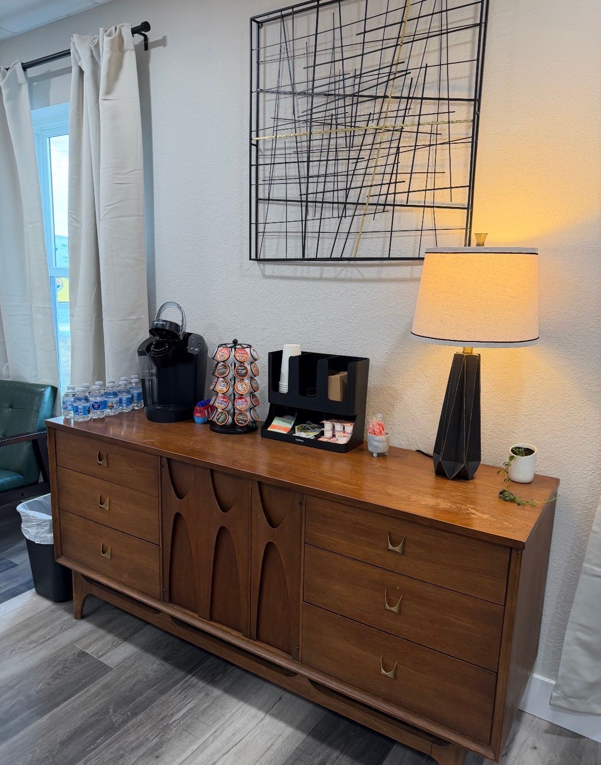 A mid-century modern wooden dresser serves as a coffee station with a machine, accessories, and a lamp under wall art.