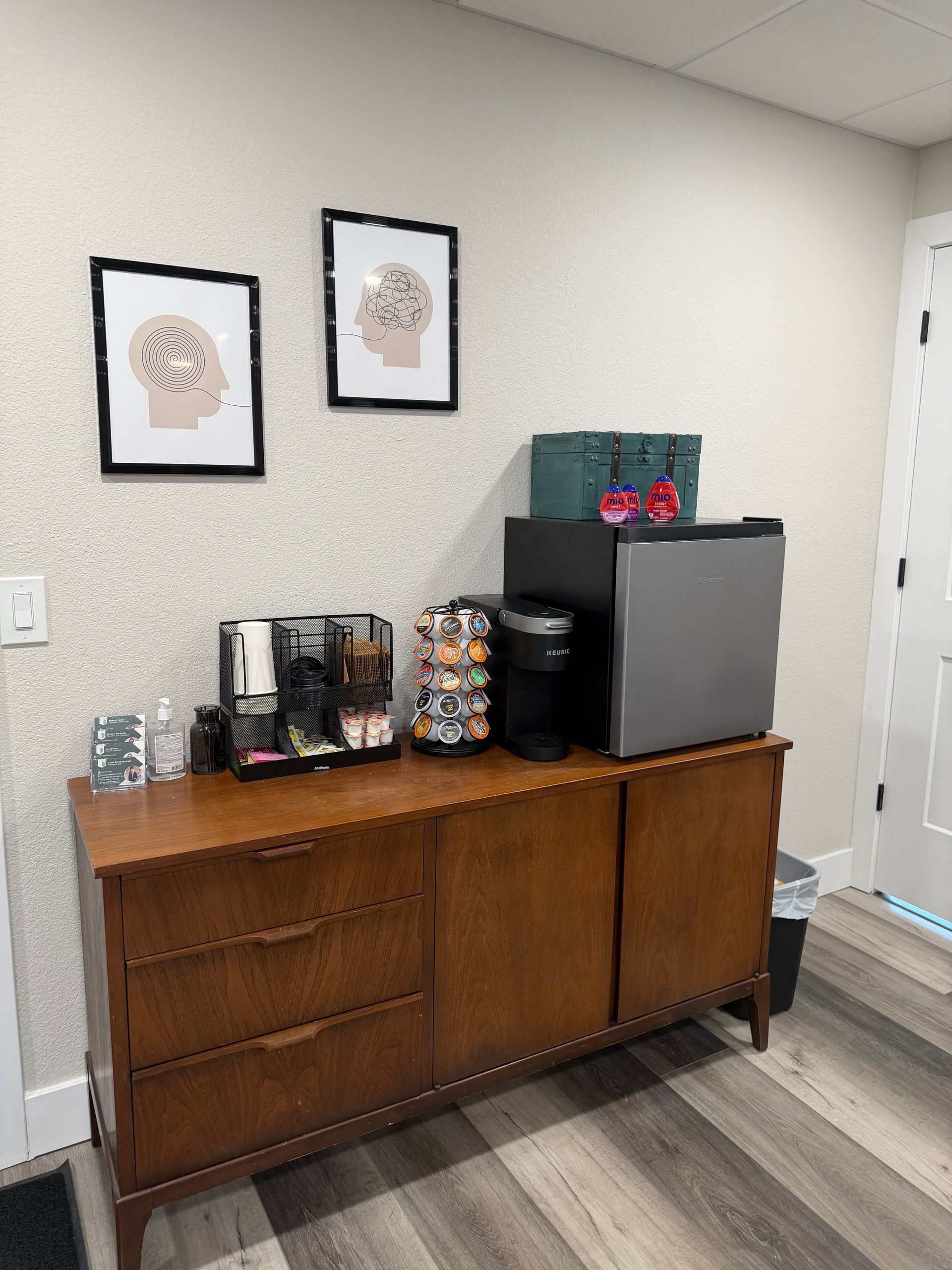 A wooden cabinet holding a coffee machine, a pod organizer, and a small refrigerator, with two brain art prints above.