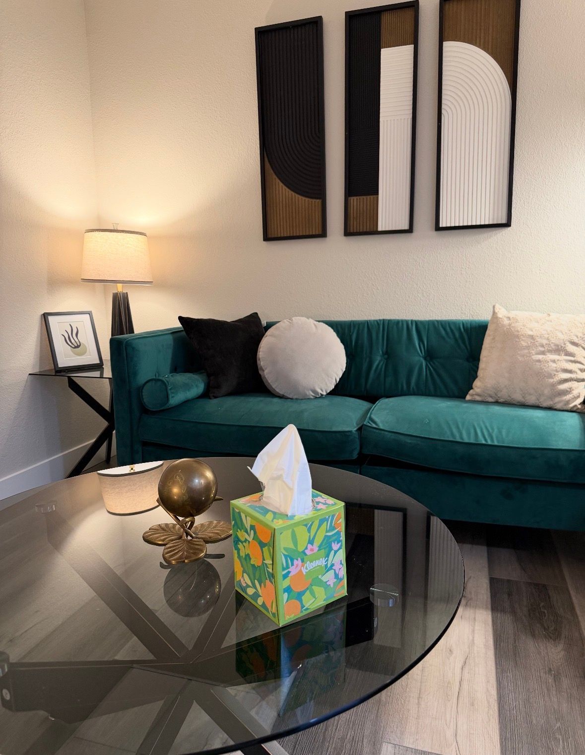 A teal sofa with patterned pillows sits before a glass coffee table with a globe and a floral tissue box in a living room.