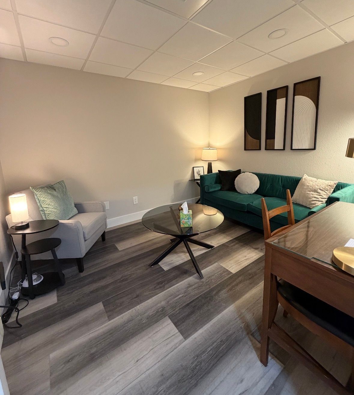 A cozy, modern office interior with a teal sofa, gray armchair, round glass coffee table, and wood-look laminate flooring.