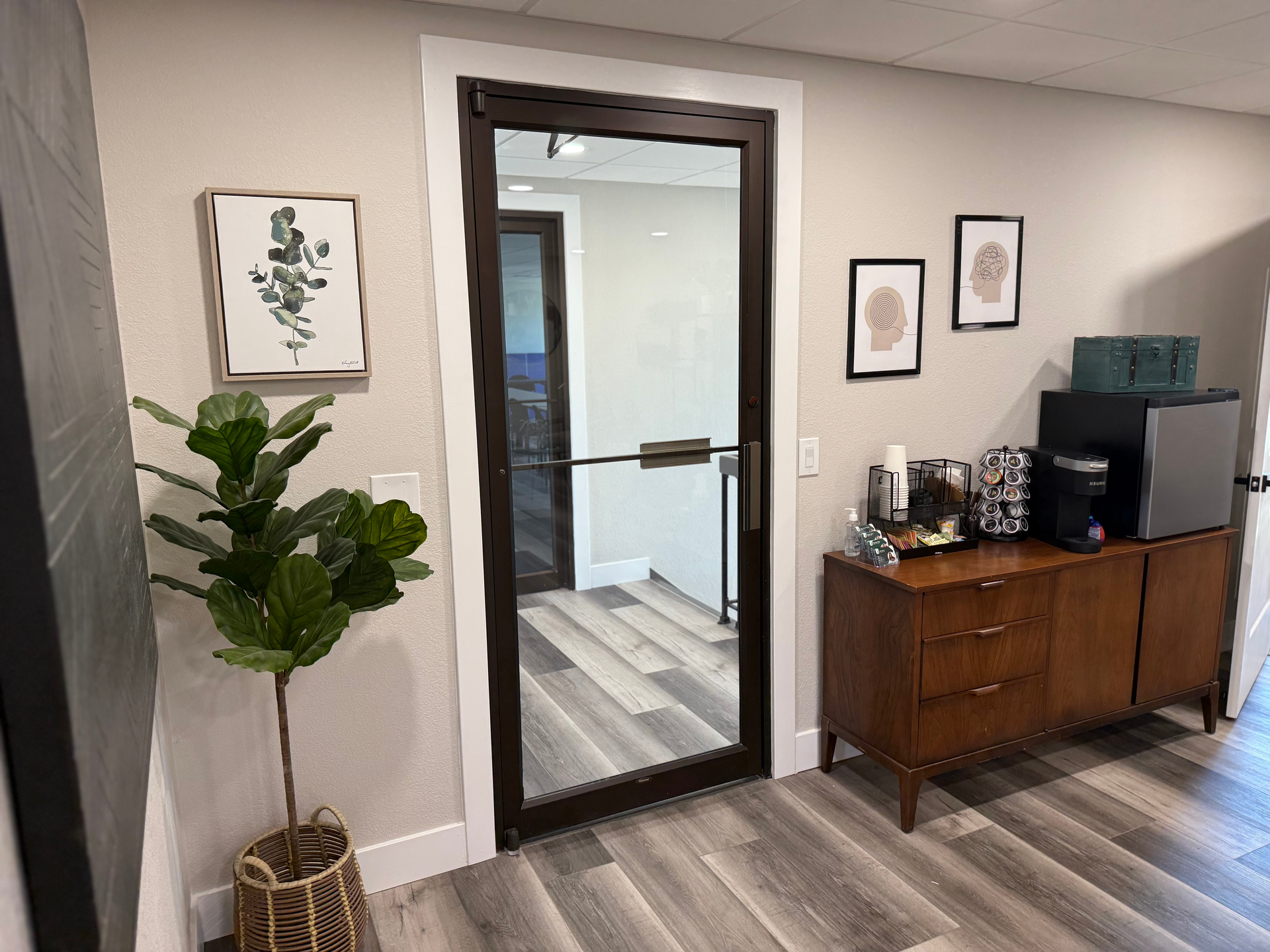 Interior with a plant, framed art, a brown cabinet, and a glass door leading to a hallway.