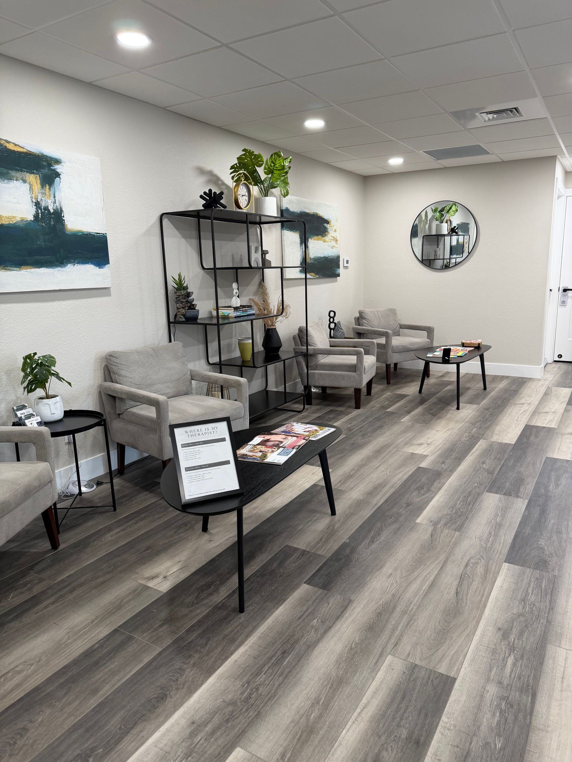 A brightly lit office waiting area with gray armchairs, a central black coffee table, open shelving, and wood-look flooring.