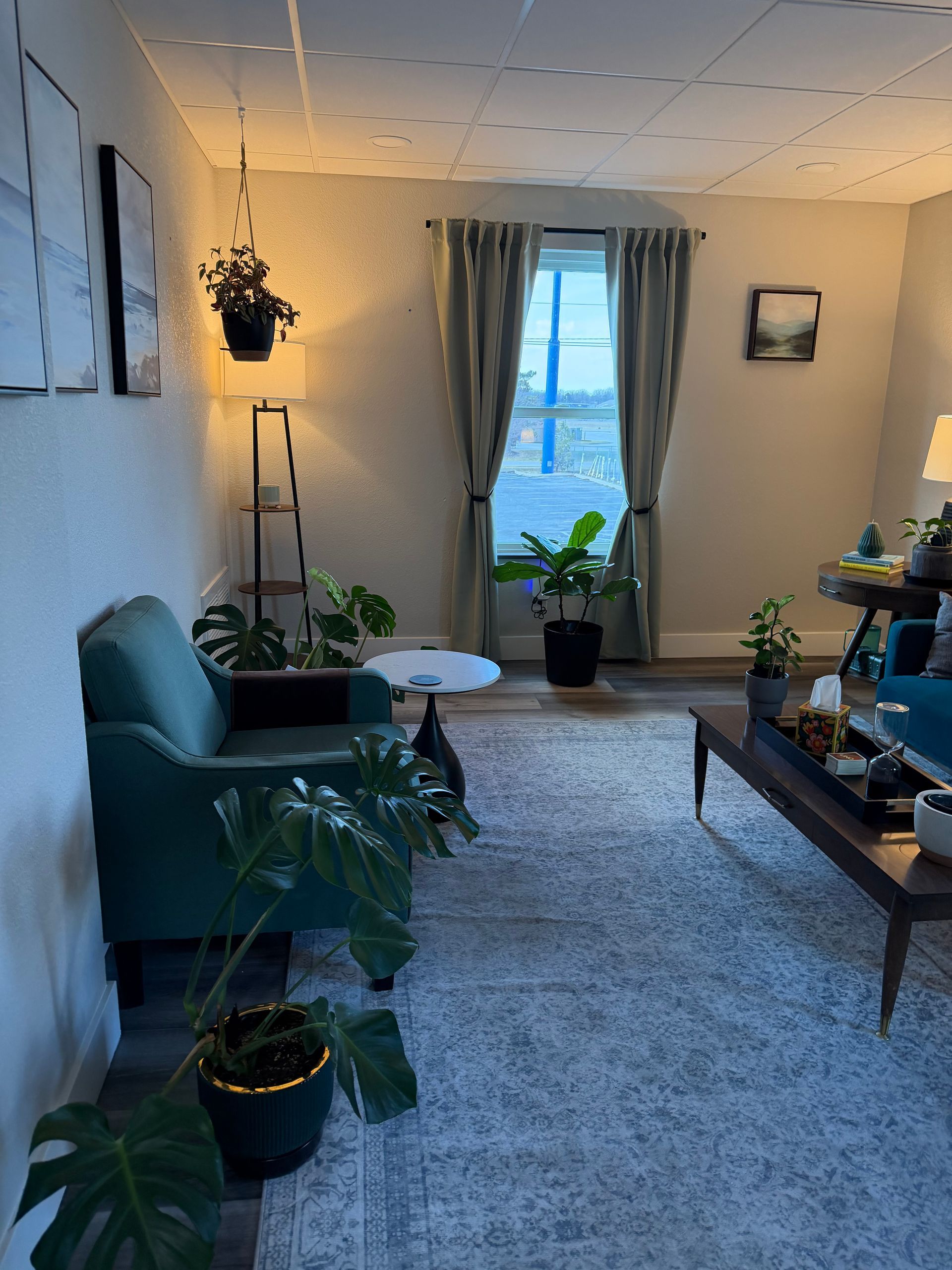 A cozy living room with a teal armchair, various indoor plants, a floor lamp, and a coffee table on a patterned rug.