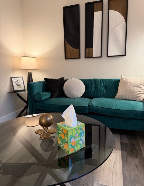 A teal velvet sofa with pillows sits in front of three abstract wall art pieces, a glass table, and a lamp.