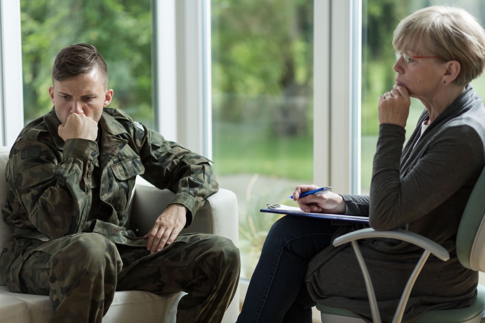 A soldier in uniform sits on a sofa, looking thoughtful, while a professional holds a clipboard in a bright office.