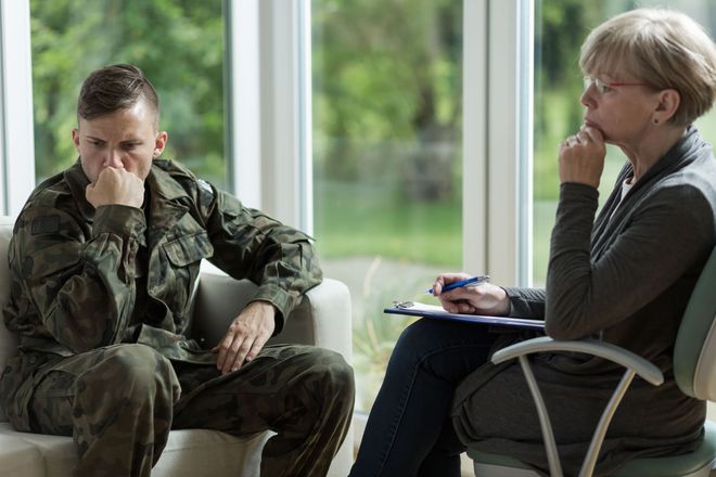 A soldier in uniform sits on a sofa, looking thoughtful, while a professional holds a clipboard in a bright office.
