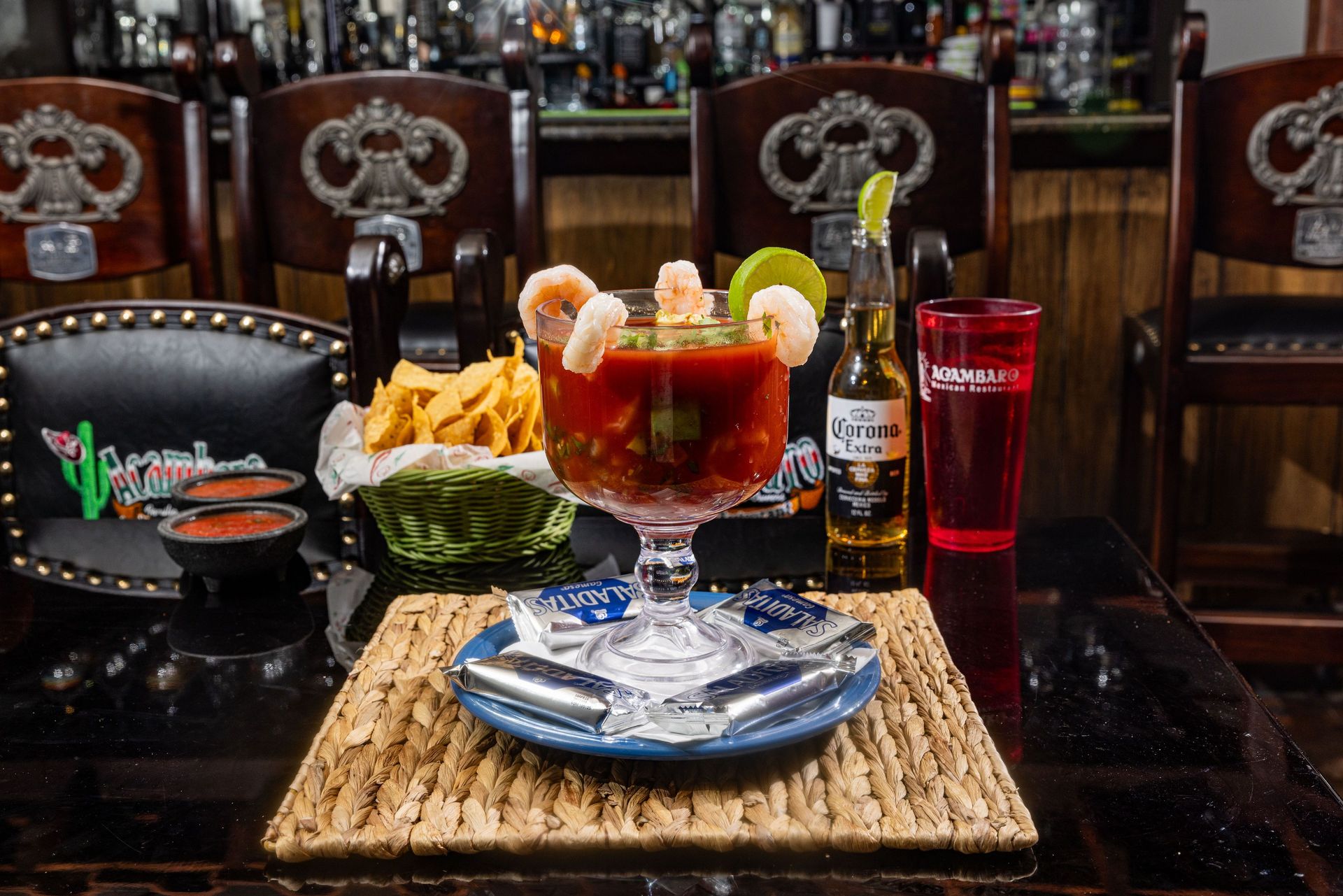 A shrimp cocktail in a large glass with chips, beer, and lime on a table.