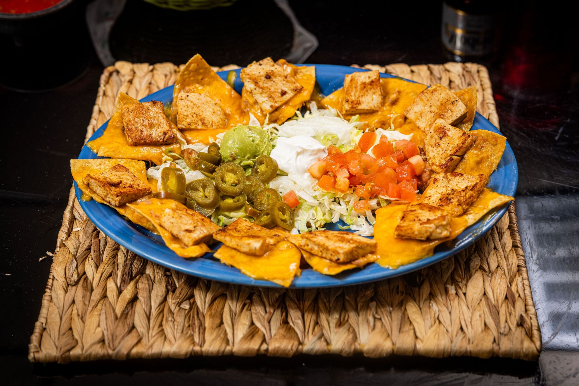 Nachos on a blue plate, topped with tofu, salsa, jalapeños, and sour cream, on a woven placemat.