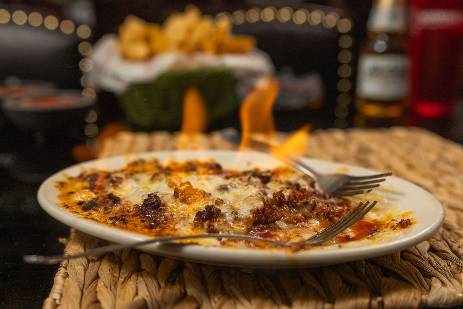 Plate of flaming, cheesy food with forks; fries and a beer bottle in the background.