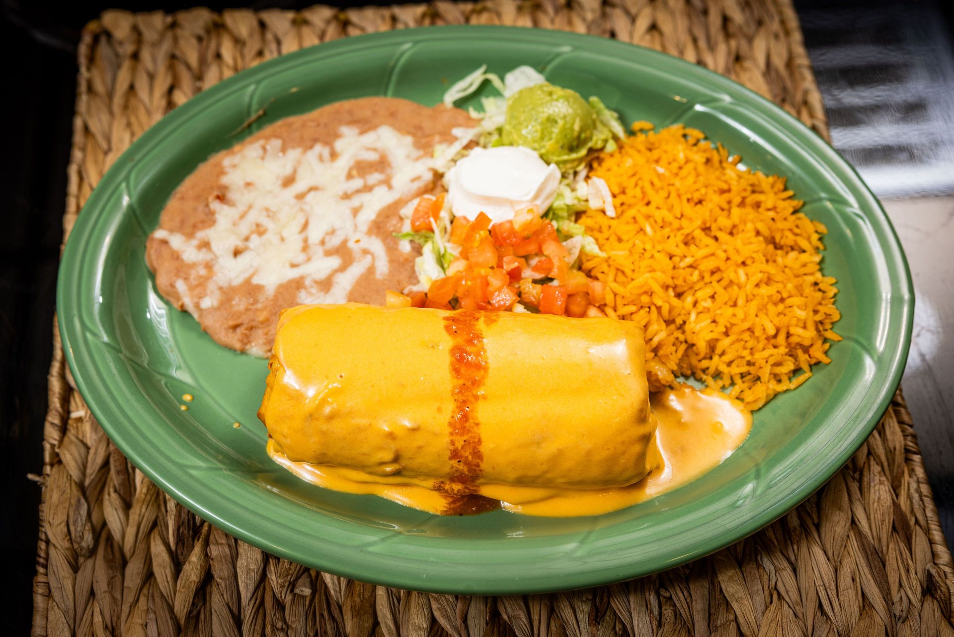 Burrito smothered in cheese sauce, with rice, beans, and garnishes on a green plate.
