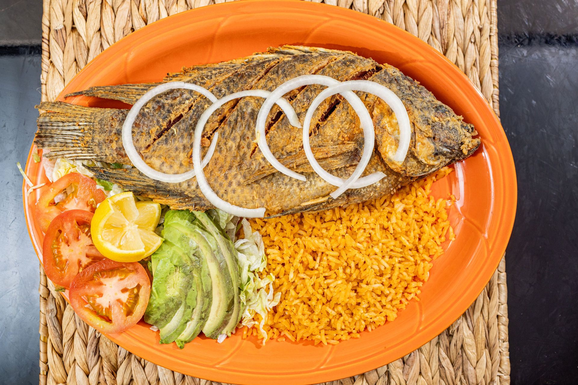 Fried fish with rice, avocado, tomato, lemon, and onion on an orange plate.