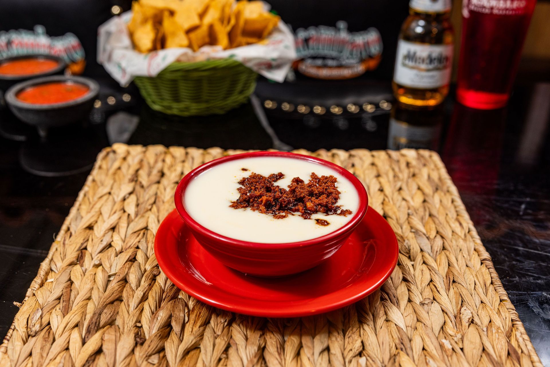 Queso in a red bowl, topped with chorizo, on a woven mat. Chips, salsa, and beer in the background.