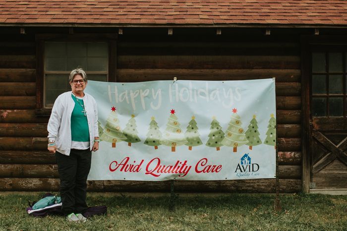 A woman is standing in front of a log cabin holding a banner.