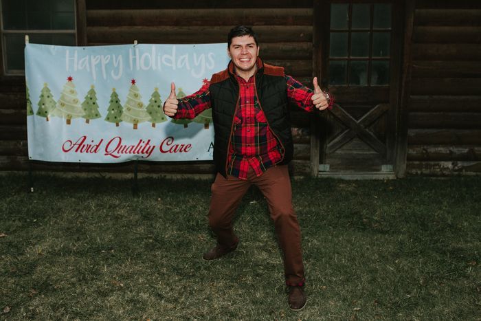 A man in a plaid shirt is giving a thumbs up in front of a sign that says happy holidays.