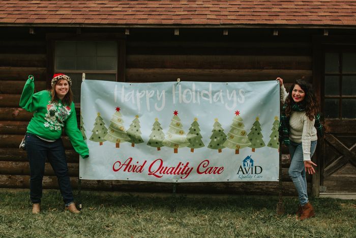 Two women are holding a christmas banner in front of a log cabin.