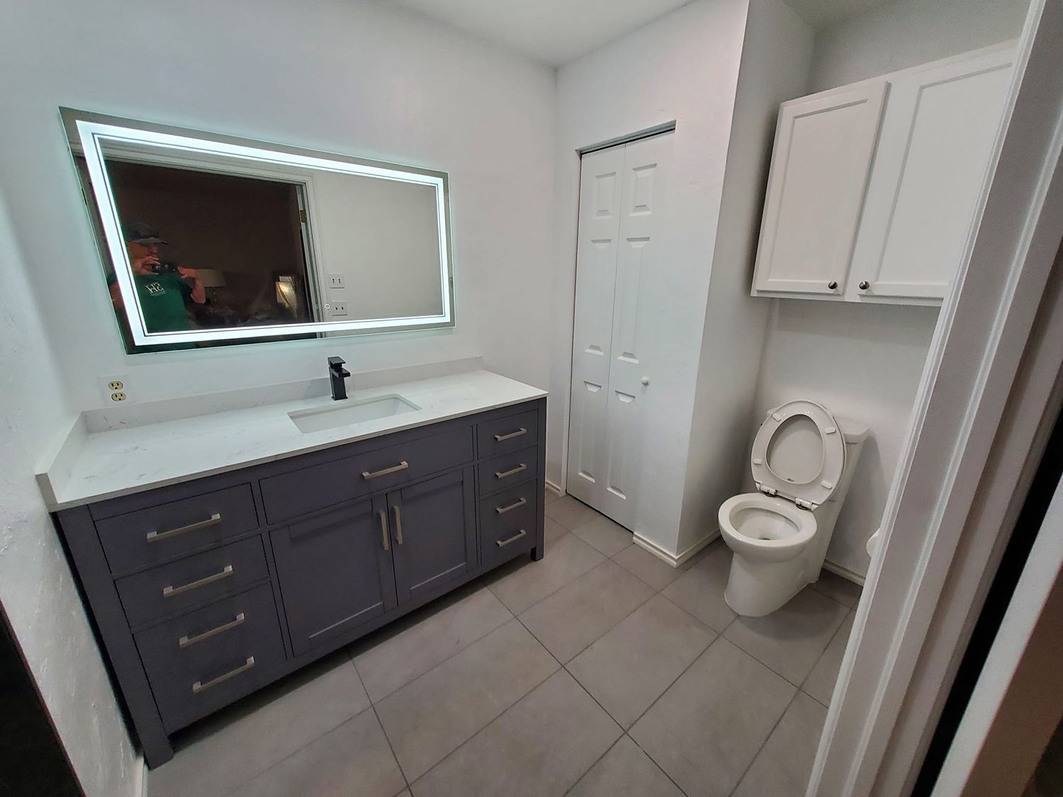 Modern bathroom with grey vanity, lighted mirror, white toilet, and gray tile floor.