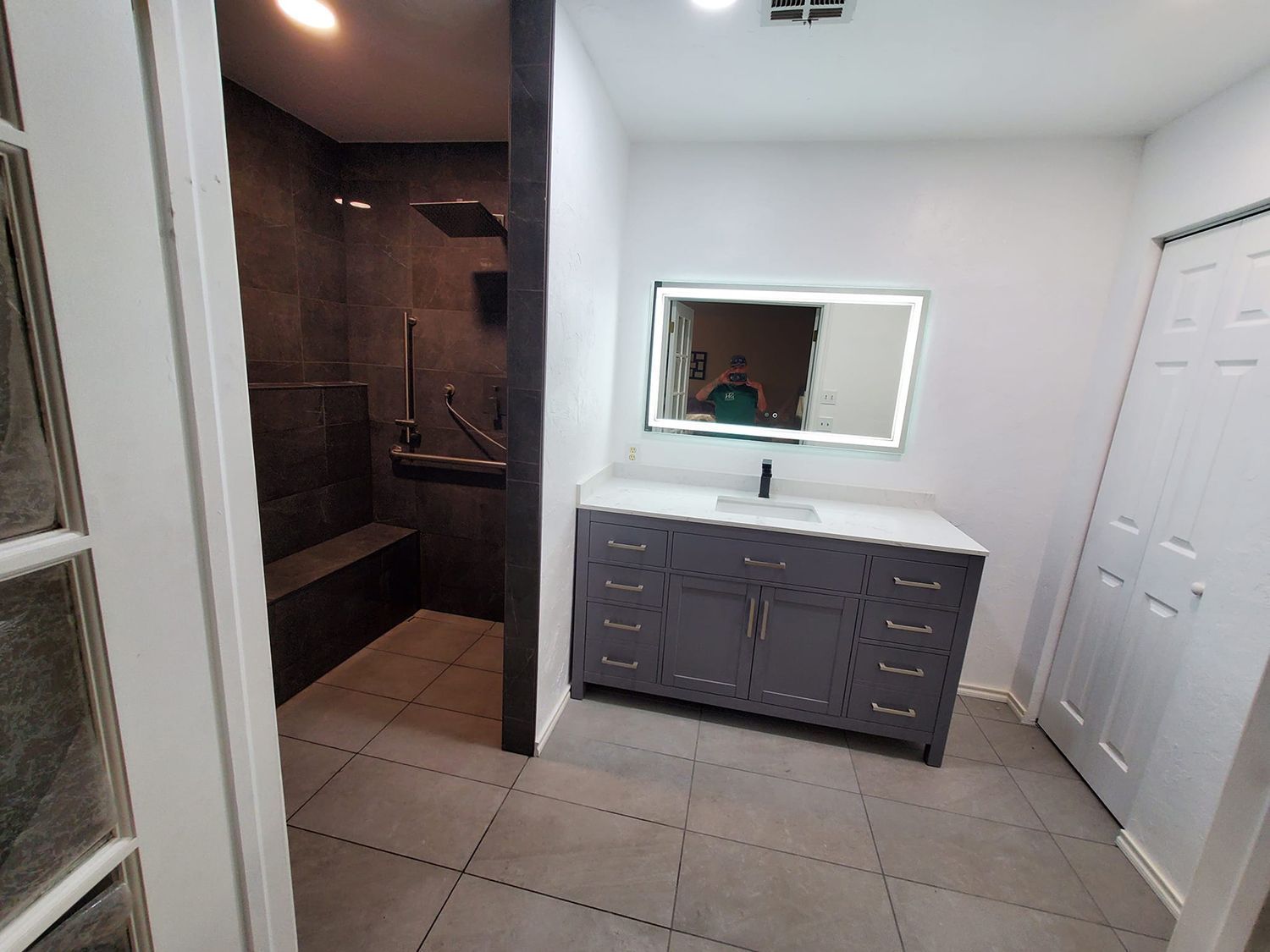 Modern bathroom with gray vanity, tiled floor, walk-in shower, and large mirror.