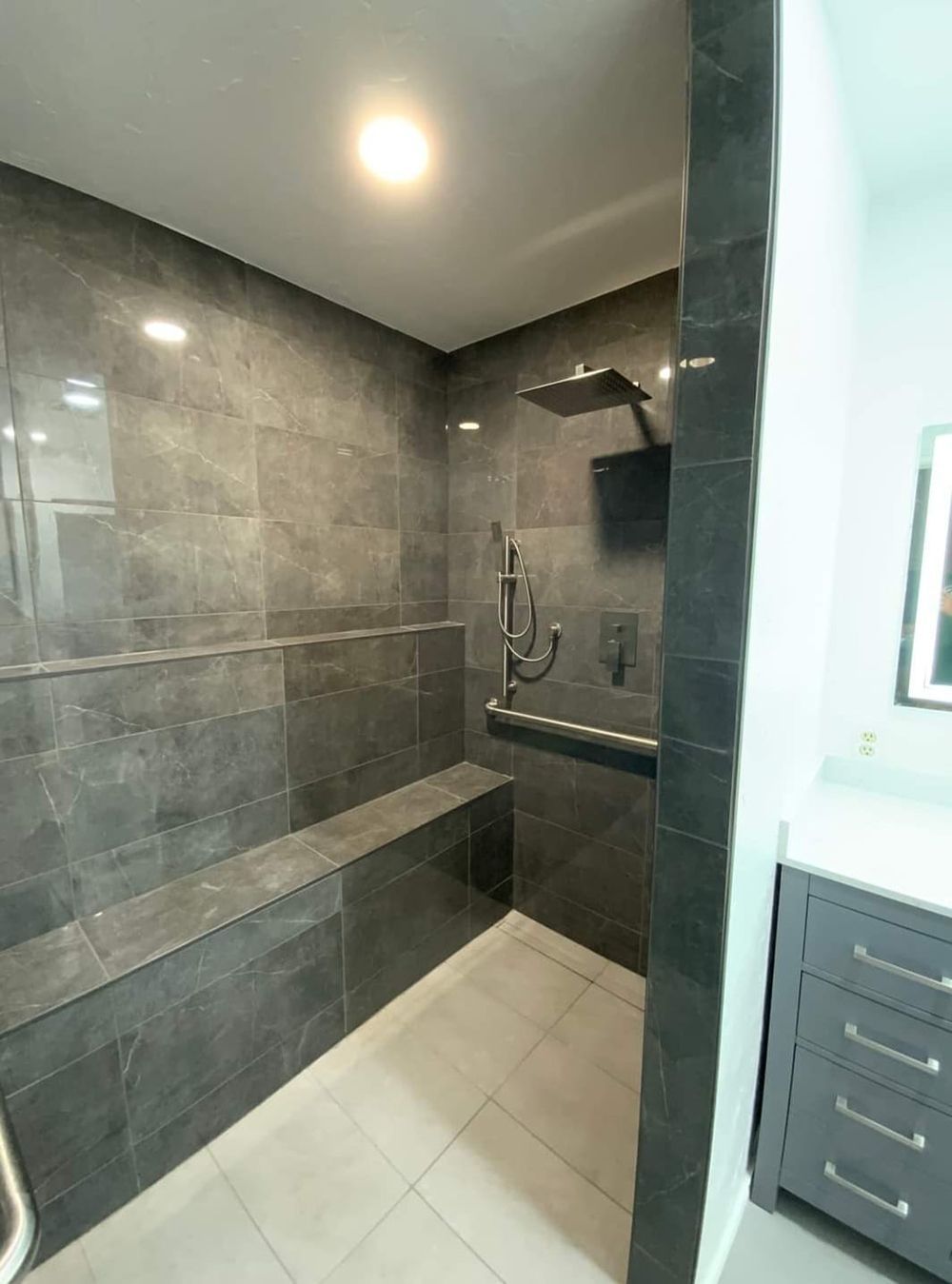 Gray tiled shower with bench, rain shower head, and handheld sprayer. White floor.