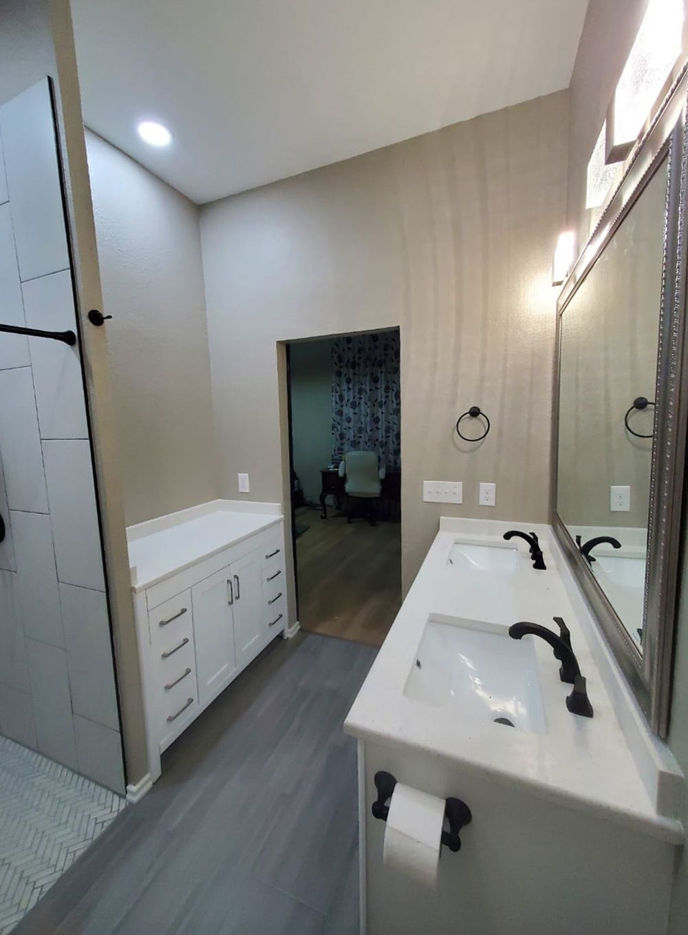 Bathroom with a double vanity, beige walls, and a view into another room.