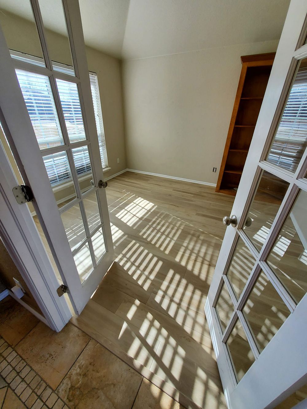 Open white French doors to a small room with wooden floor, windows with blinds, and a bookcase.