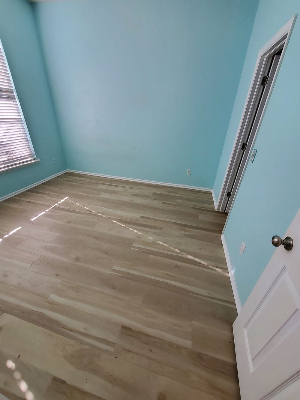 Empty room with light wood flooring, turquoise walls, and a white door and trim. Window with blinds.