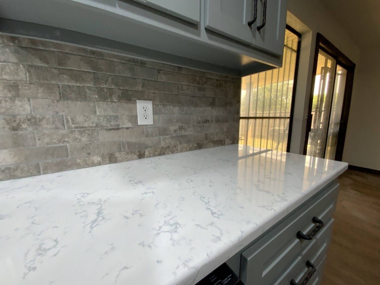 Kitchen with gray cabinets, white countertop, and stone backsplash; light streams in from a doorway.
