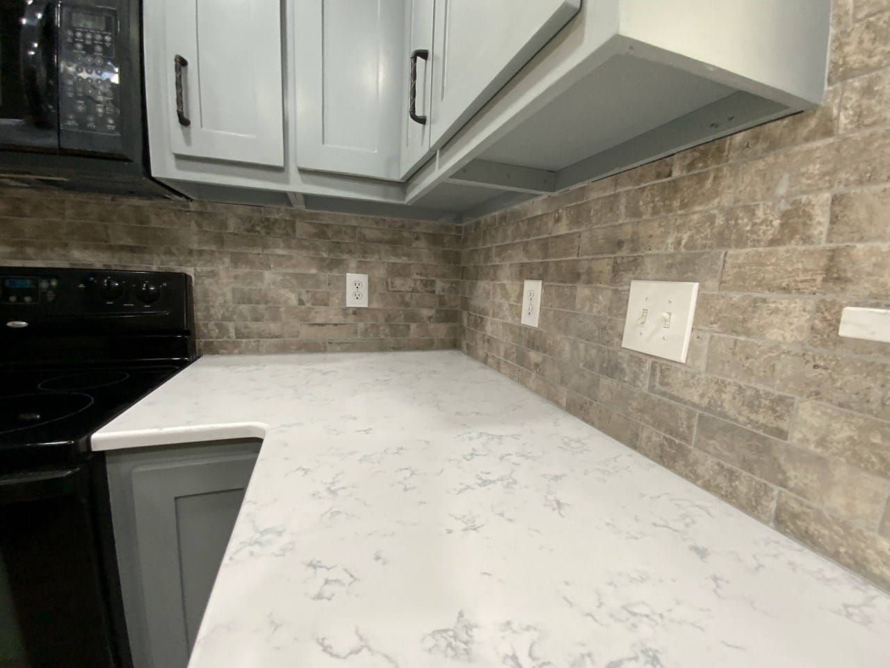 Kitchen counter with white surface, gray cabinets, and stone backsplash. Black appliances are present.