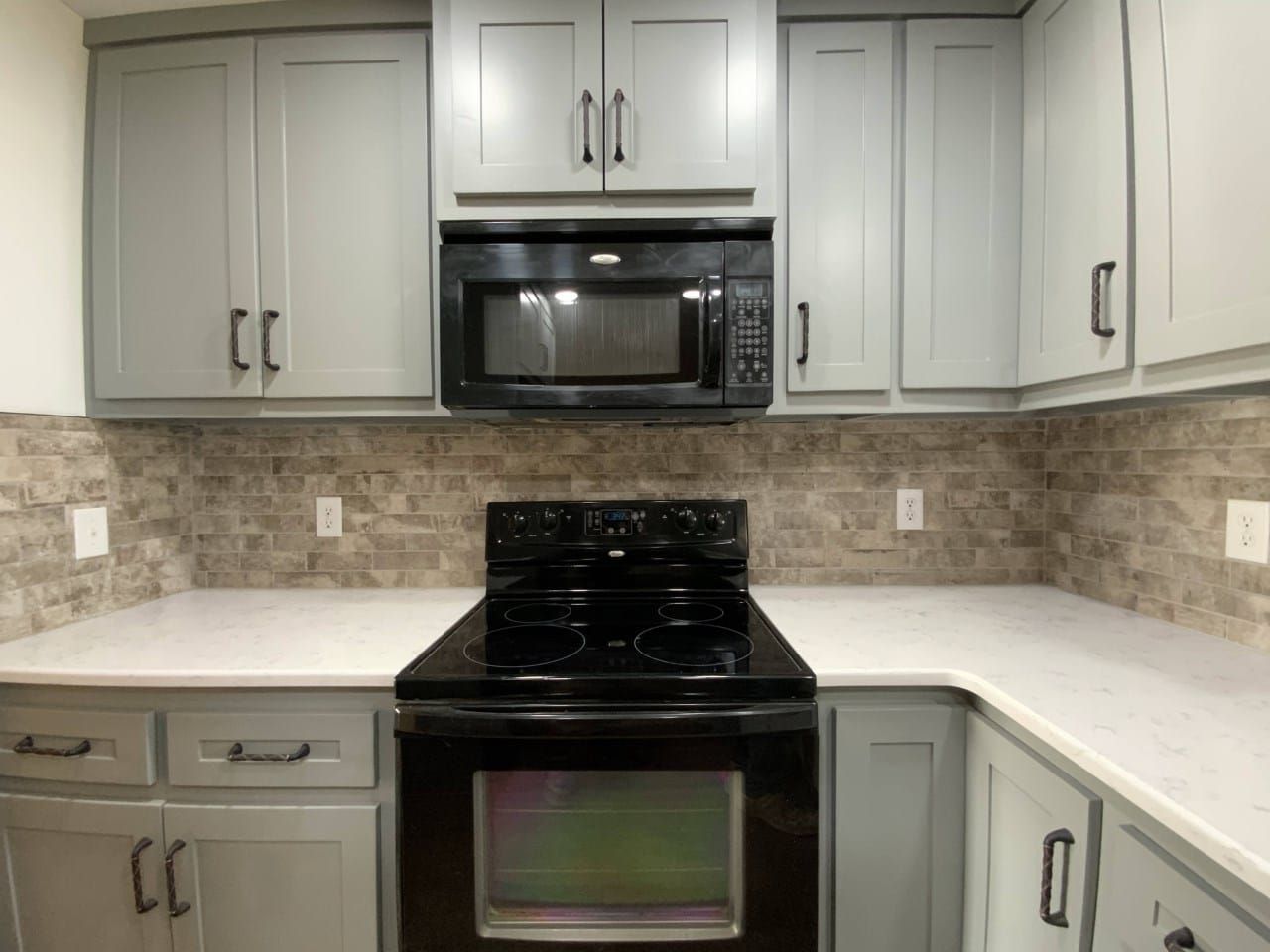 Gray kitchen cabinets with a black microwave and oven, white countertops, and brick-style backsplash.
